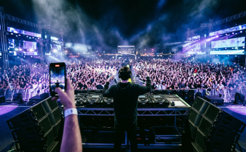 AI-generated editorial photo of a DJ performing on a mainstage festival stage at night, seen from behind, facing a massive crowd with dramatic colored lighting